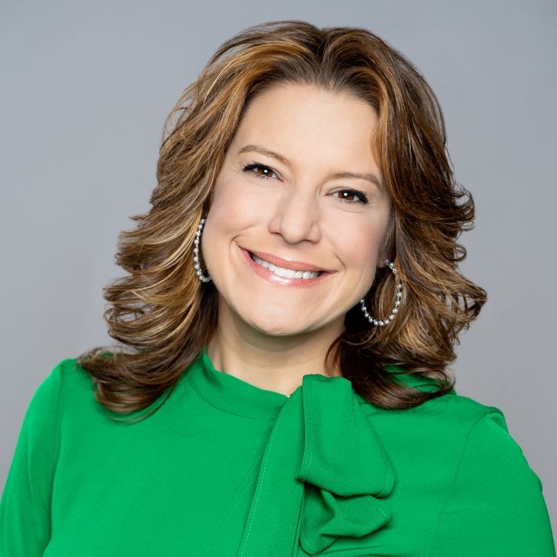 A woman with shoulder-length wavy hair wearing a bright green top with a bow detail and pearl earrings, smiling against a neutral background.