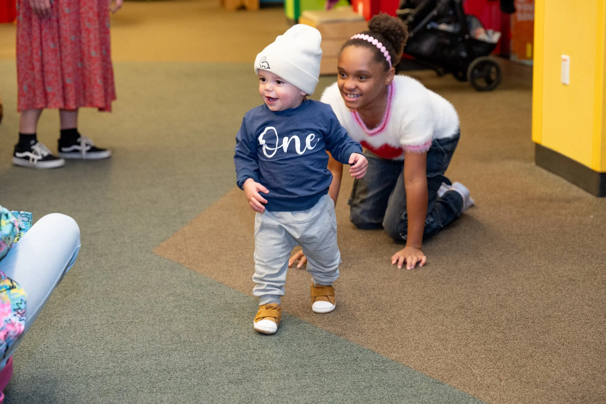 A toddler wearing a white beanie, navy blue shirt with 'One' written on it, light gray pants, and brown shoes, walking on a carpeted floor. A smiling woman is kneeling behind the toddler, dressed in a white fuzzy sweater with pink accents and dark pants. There are others in the background and the setting appears to be indoors.