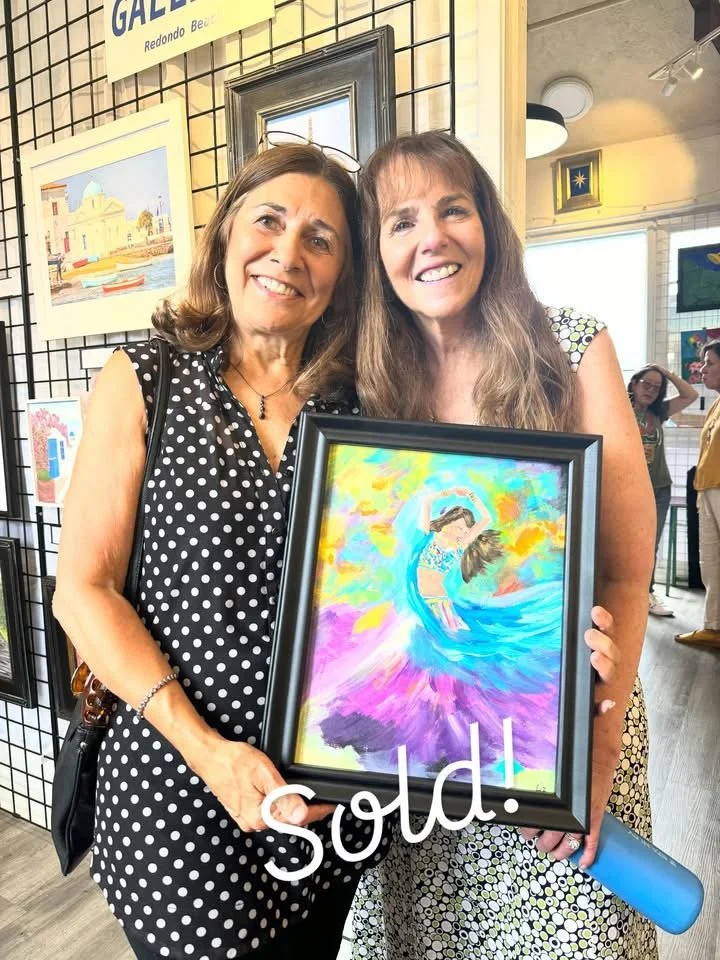 Two women smiling, holding a framed colorful painting of a woman dancing in a blue dress with rainbow background, at an art gallery.