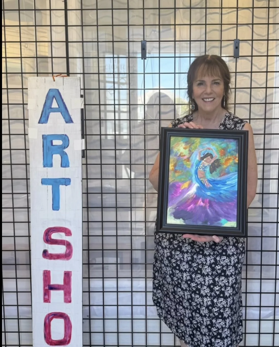 A woman holding a colorful painting of a dancer in a flowing dress and dancing pose, standing next to a sign spelling 'ARTS' on a wire grid backdrop.