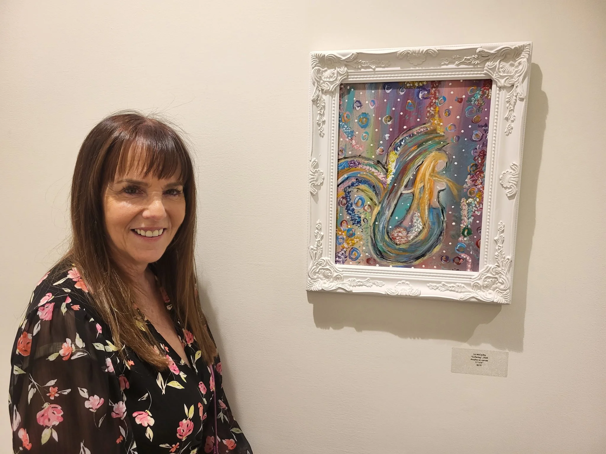 A woman with light brown hair and a floral blouse is smiling in front of a colorful abstract painting framed in white. The painting is titled 'Surface' by artist Liz McCarthy, created in 2025, and is displayed at an art gallery.