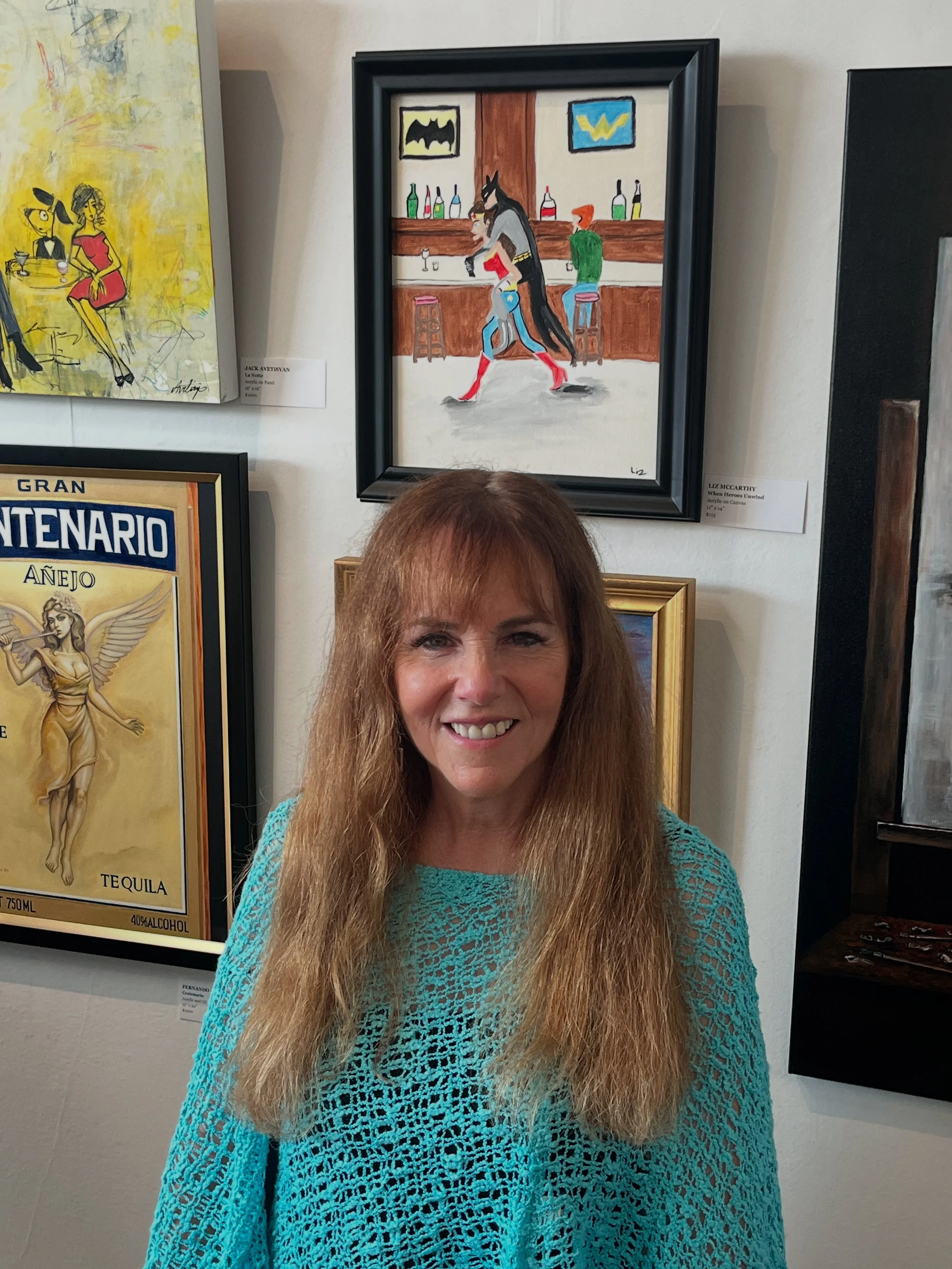 A woman with long, light brown hair smiling at the camera, wearing a turquoise crocheted top. She is standing in front of framed paintings on a white wall that include a colorful cartoon scene of a bar with characters and portraits.