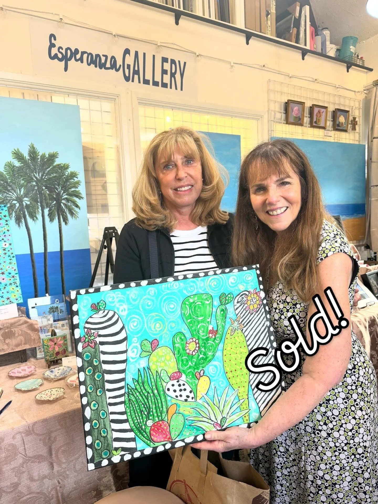 Two women at an art gallery, smiling, holding a colorful abstract painting of cacti and flowers. Behind them are other artworks, including paintings of palm trees and beach scenes, on display at the Esperanza Gallery.
