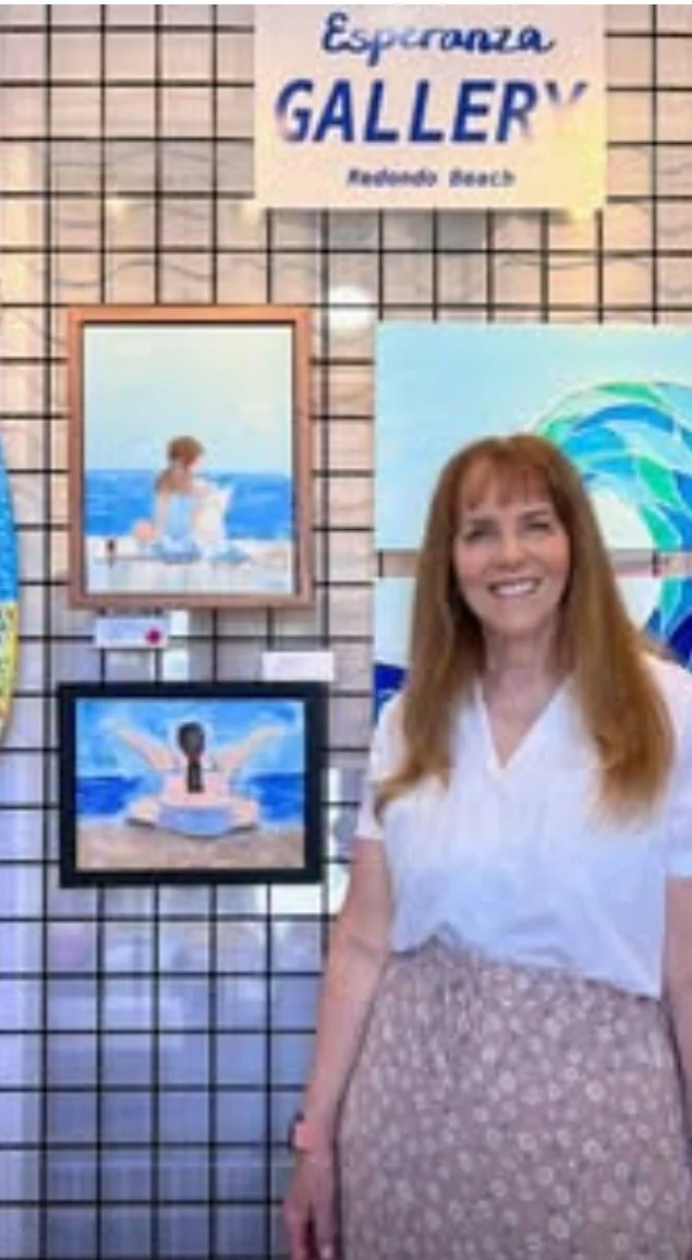 A woman with red hair smiling in front of artwork at the Esperanza Gallery, with framed paintings of ocean scenes and a sign above that reads 'Esperanza GALLERY.'