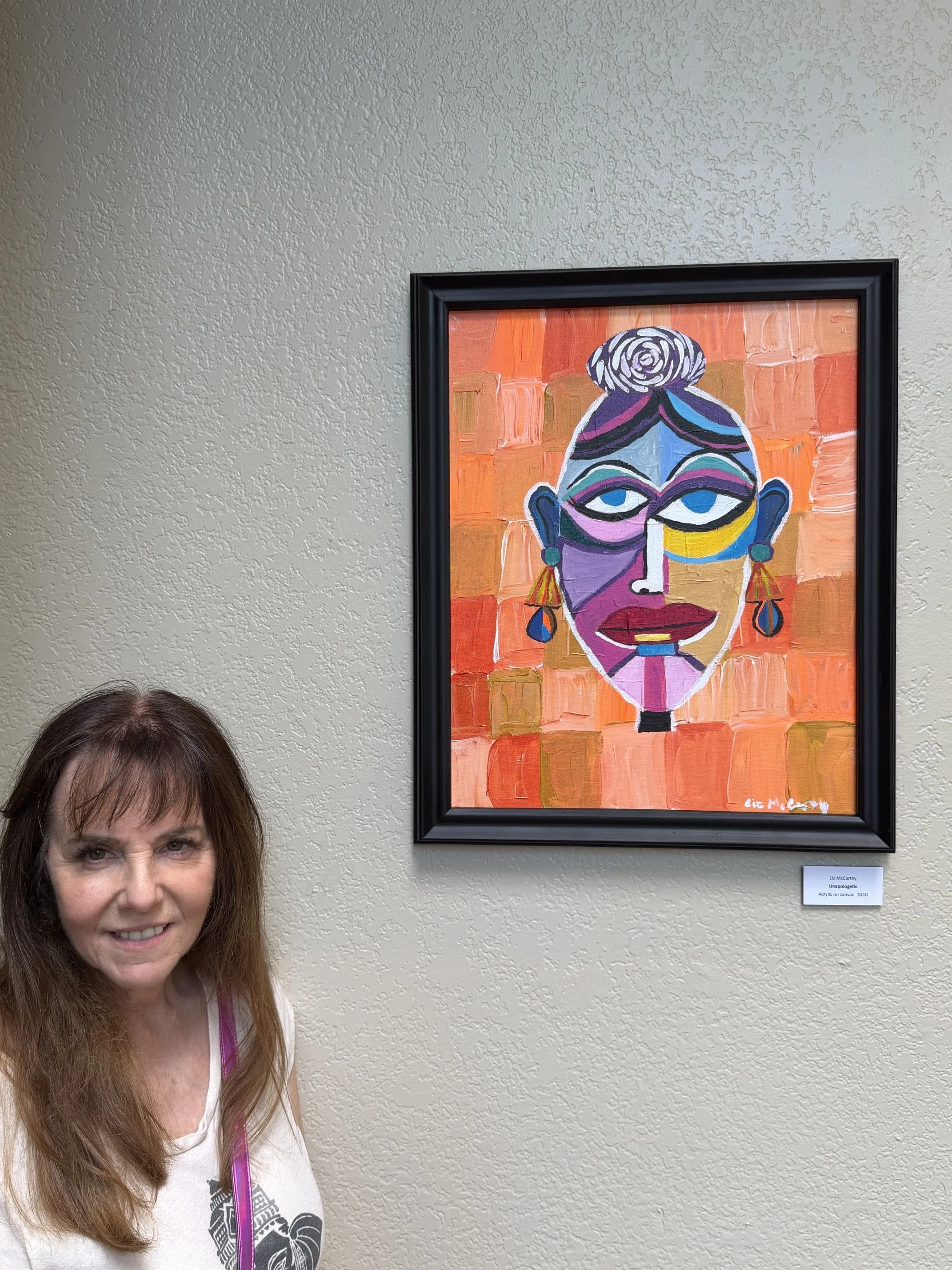 A woman with long hair smiling in front of a colorful abstract portrait painting of a woman with an orange background.