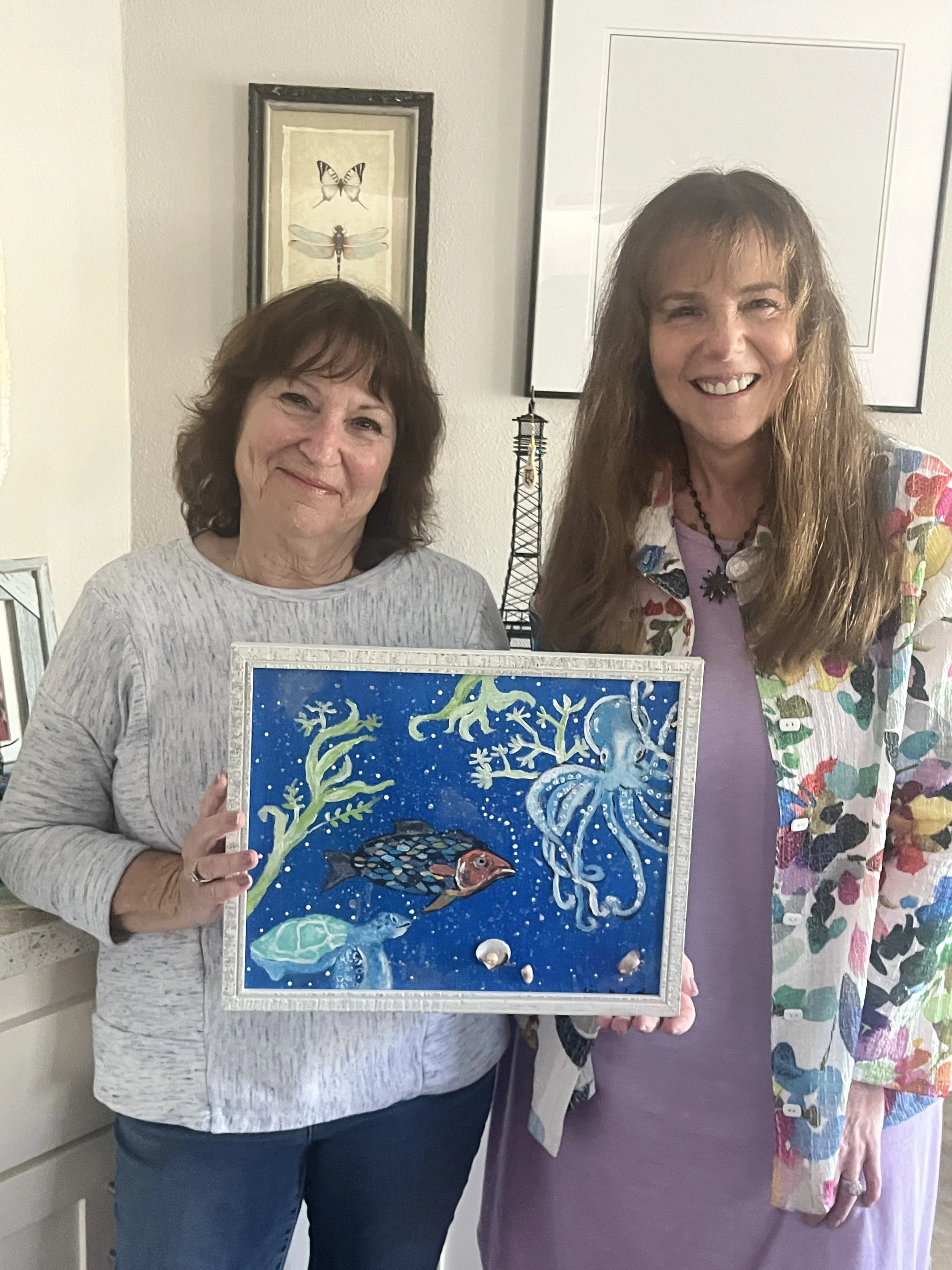 Two women smiling, one holding a colorful painting of an underwater scene with fish, coral, and seaweed.
