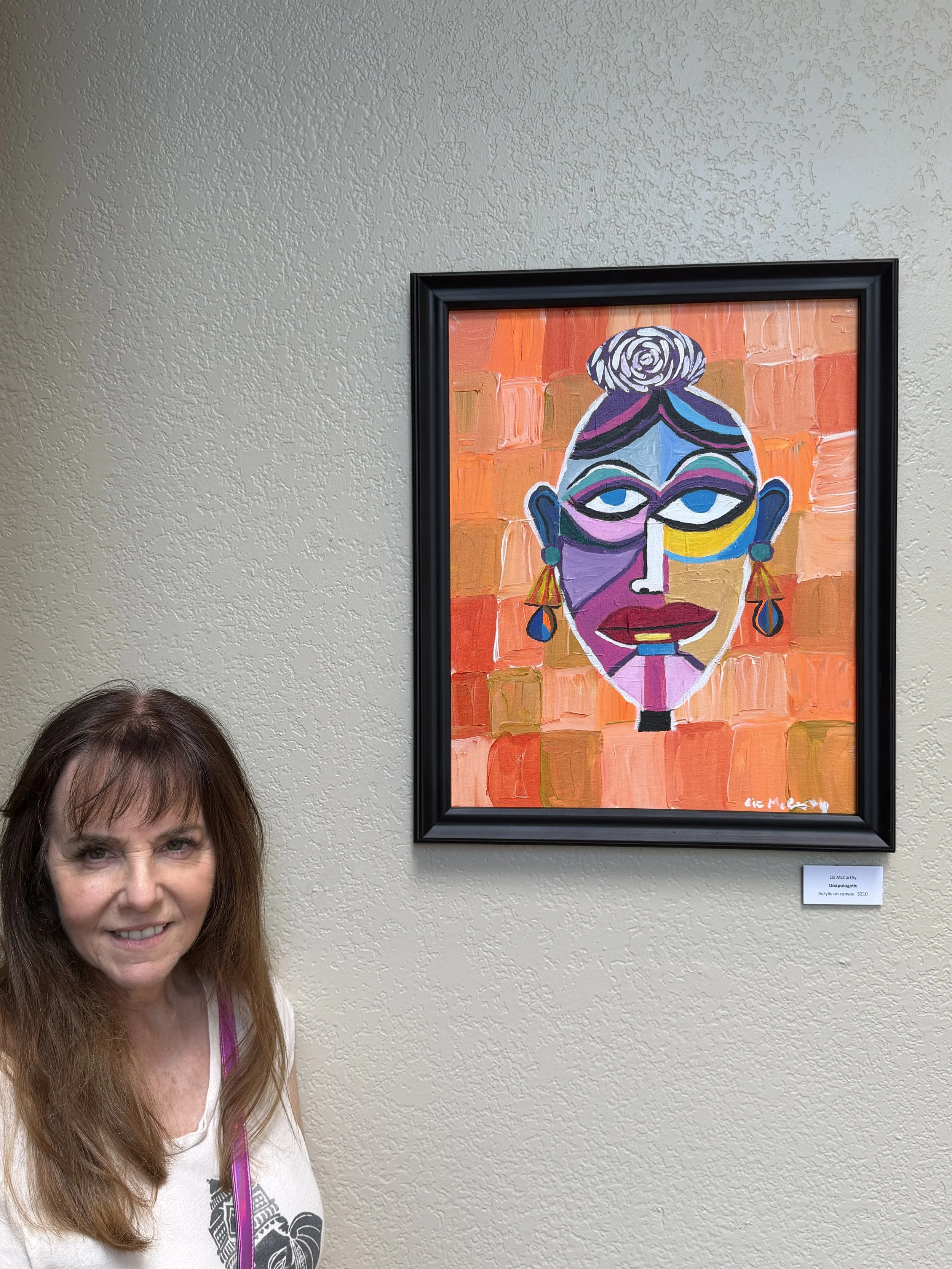 Portrait of a woman with long brown hair and a white shirt, standing next to an abstract, colorful painting of a face with bold lines and geometric shapes, hung on a textured beige wall