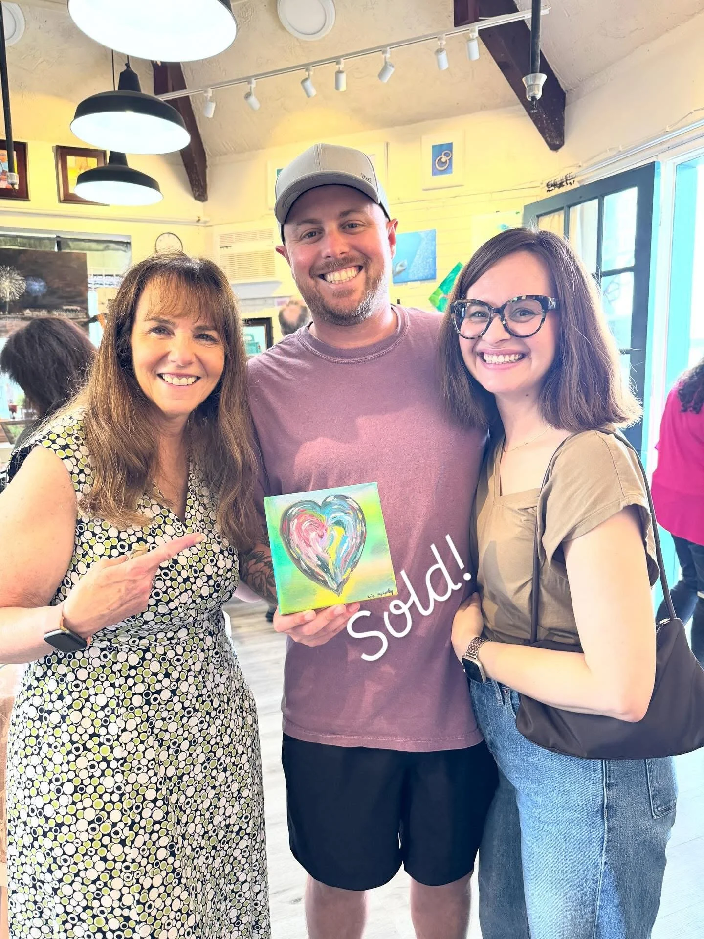 Three people standing together indoors, smiling, with a man in the middle holding a colorful artwork featuring a heart and the word 'Sold!' on it. The woman on the left is wearing a patterned dress, the man is wearing a cap and a casual t-shirt, and the woman on the right is wearing glasses and a beige top. The background shows art on the walls, a window, and hanging lights.