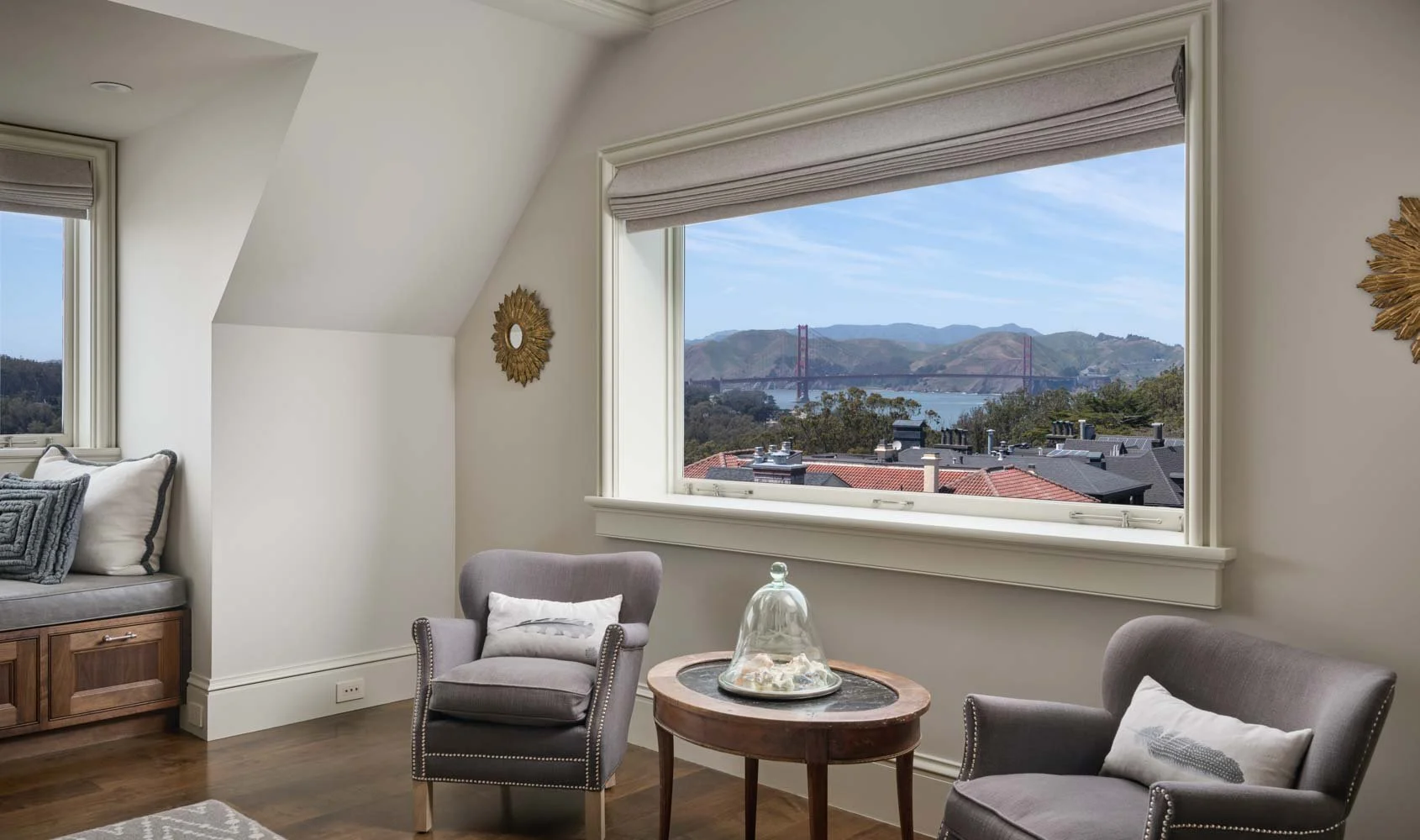 A cozy living room with two gray armchairs and a wooden side table with a glass cloche decor, large window showing a view of hills, water, and the Golden Gate Bridge in San Francisco, with a glimpse of roof tops outside.