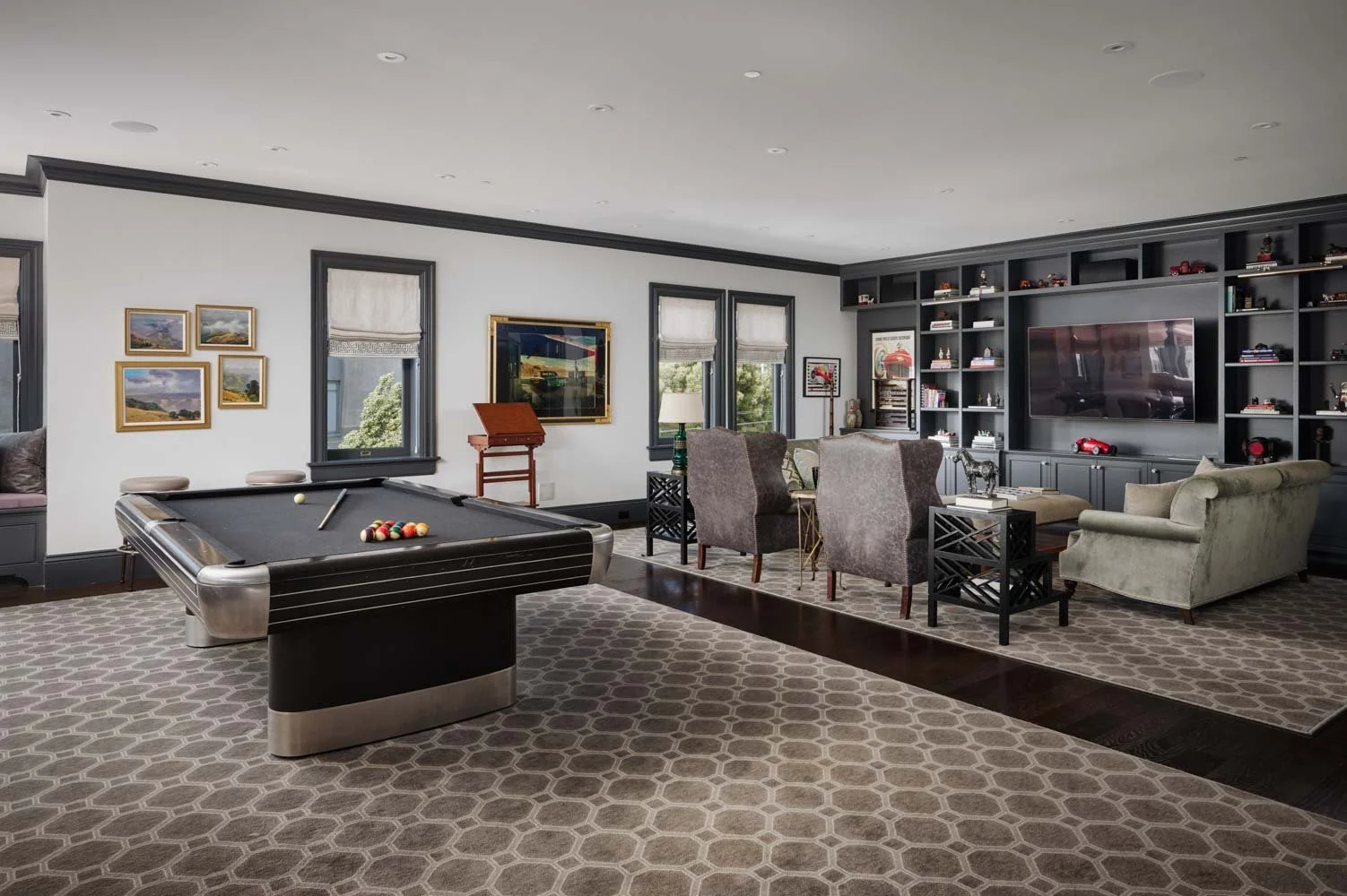 A spacious living room with a pool table, a large flat-screen TV, and built-in bookshelves filled with books and decorative items. The room features light-colored walls, dark trim, and a patterned area rug.