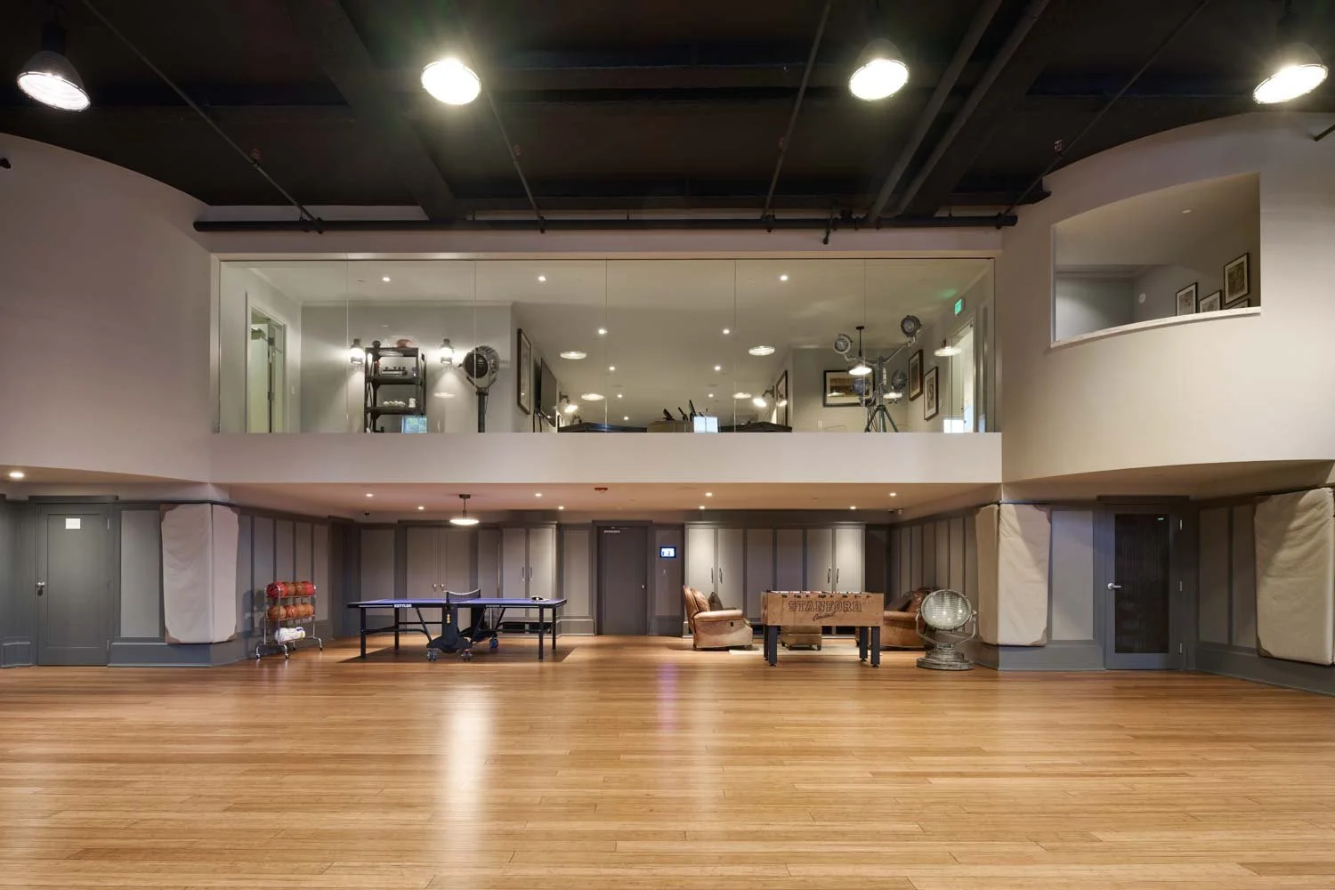 A spacious indoor recreational area with a wooden floor, a ping pong table, seating area, and a second-floor balcony with glass railing overlooking the space. The ceiling has black exposed beams and recessed lighting.