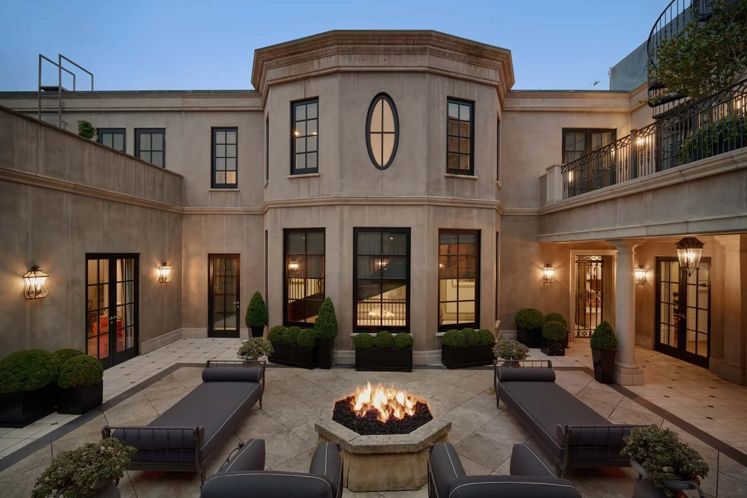 Luxury townhouse courtyard with fire pit, outdoor seating, potted plants, and exterior lighting at dusk.