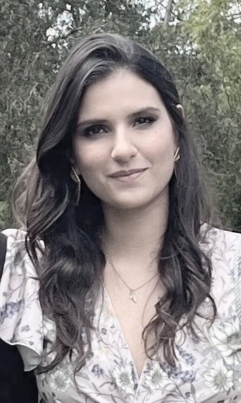 A young woman with long dark hair, wearing a light-colored floral top, standing outdoors with trees in the background