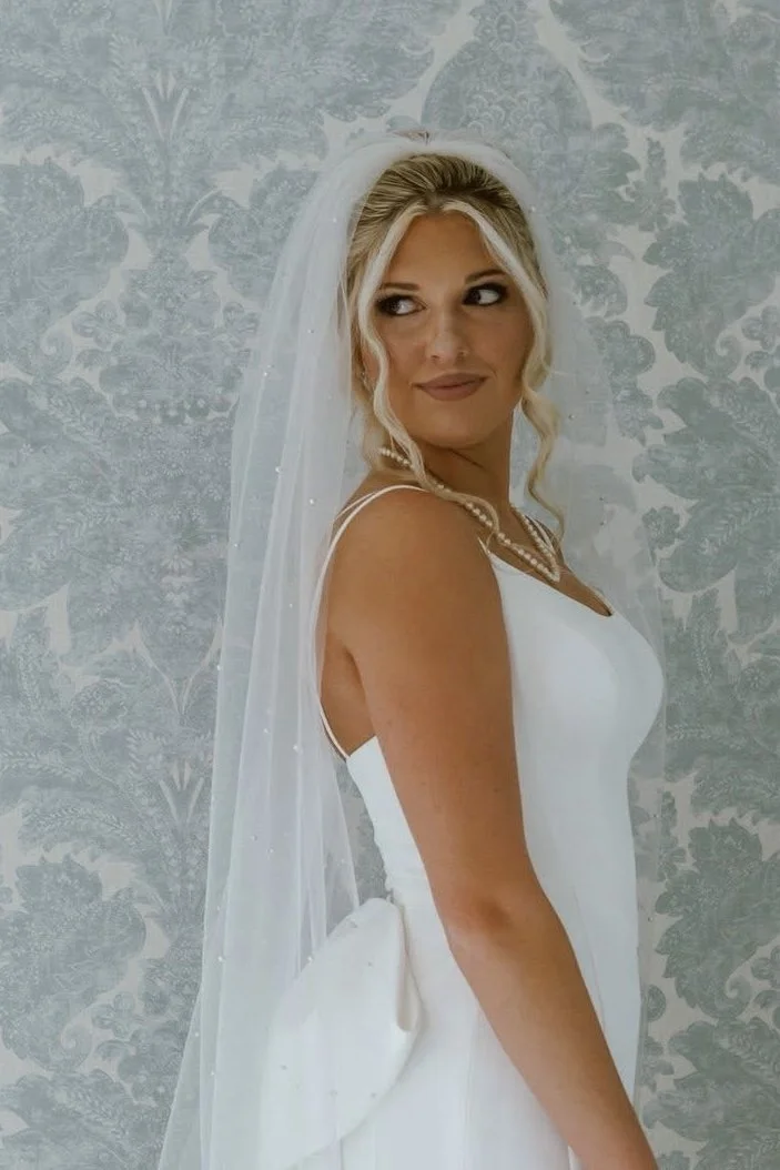 A bride wearing a white dress with a sheer veil, standing against a patterned gray background, with blonde hair styled in loose waves and a pearl necklace.