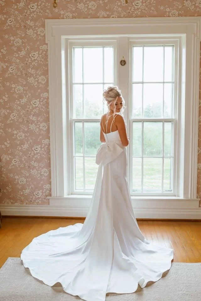 A bride in a white wedding gown with a long train standing in front of a large window with white frames, inside a room with floral wallpaper and hardwood floors.