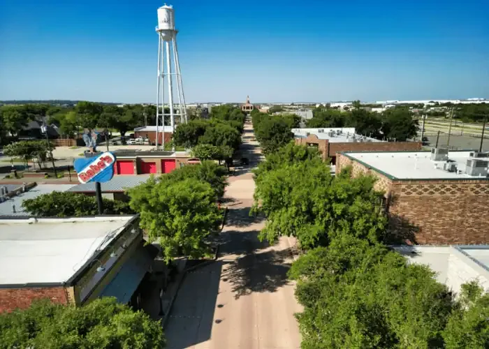 Aerial view of a small town with a sidewalk lined by trees, a water tower, and various storefront buildings under a clear blue sky.