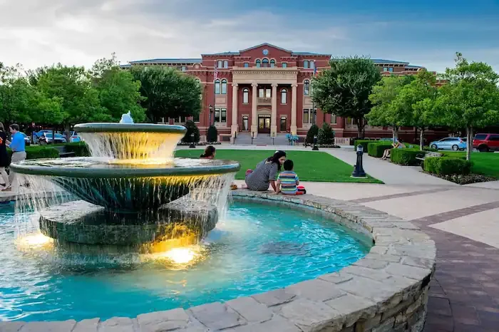 Public park with a fountain, trees, and a large red brick building in the background, with people sitting near the fountain.