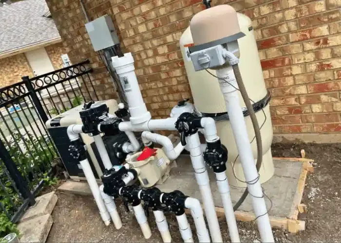 Pool filter and pump system with white PVC pipes and multiport valve outside a brick house.