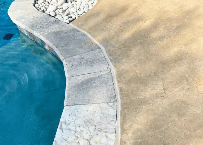 Close-up of a swimming pool's edge with a stone border, concrete decking, and decorative white rocks.