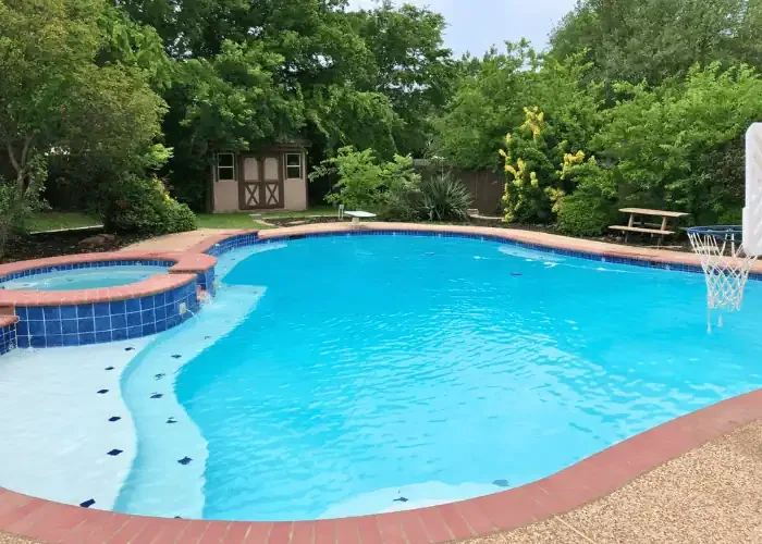 In-ground swimming pool with a hot tub, surrounded by trees and greenery, with a basketball hoop at the pool's edge.