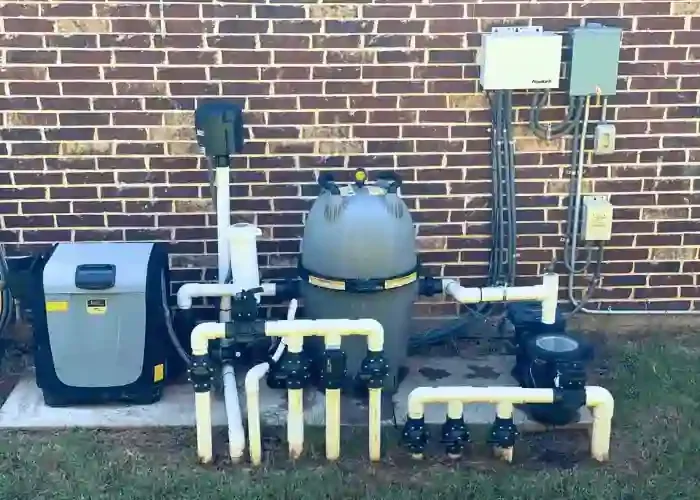 An outdoor pool filtration system with pump, filter, and piping mounted against a brick wall.