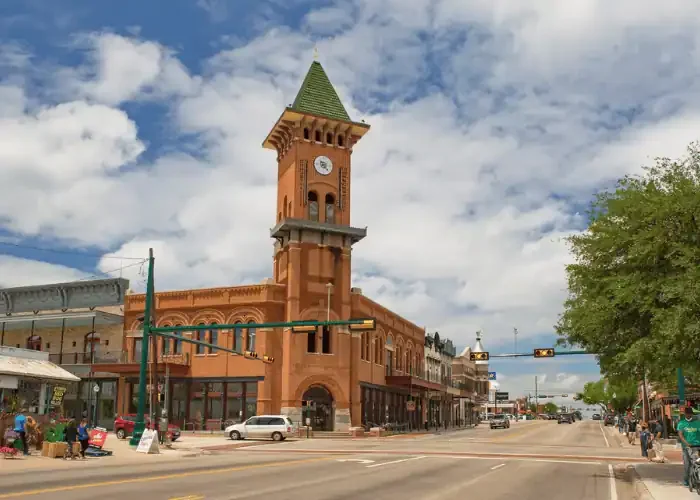Historic brick building with a tall clock tower and green roof, located on a busy town street with cars and pedestrians, under a partly cloudy sky.
