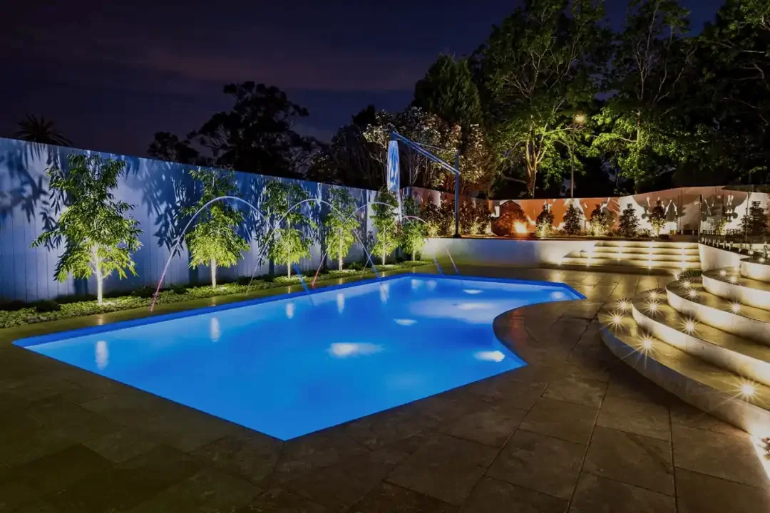 Nighttime backyard scene with illuminated swimming pool, surround sound landscaping trees and steps, and a curved patio area.