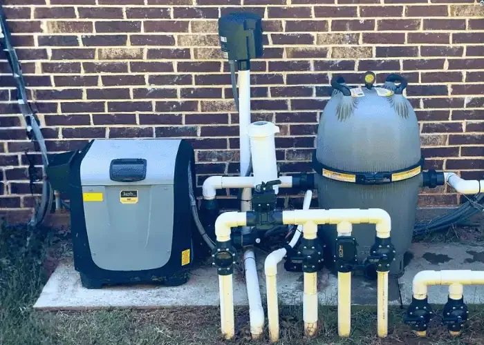 An above-ground swimming pool filter and pumping system installed next to a brick wall, with white pipes and black valves connected to the filter unit.