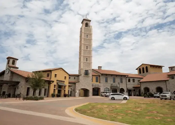A European-style building with a tall clock tower against a cloudy sky, a parking lot with cars, and a grassy area in front.