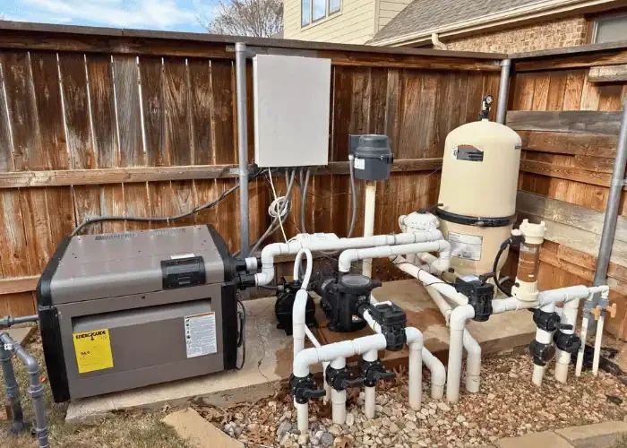 In-ground pool or spa pump and filtration system with pipes, pumps, and a control box outside on a concrete slab surrounded by gravel, next to a wooden fence.