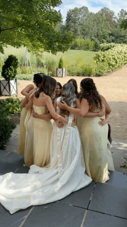 A bride in a white wedding dress hugging five bridesmaids in yellow dresses outdoors on a sunny day.