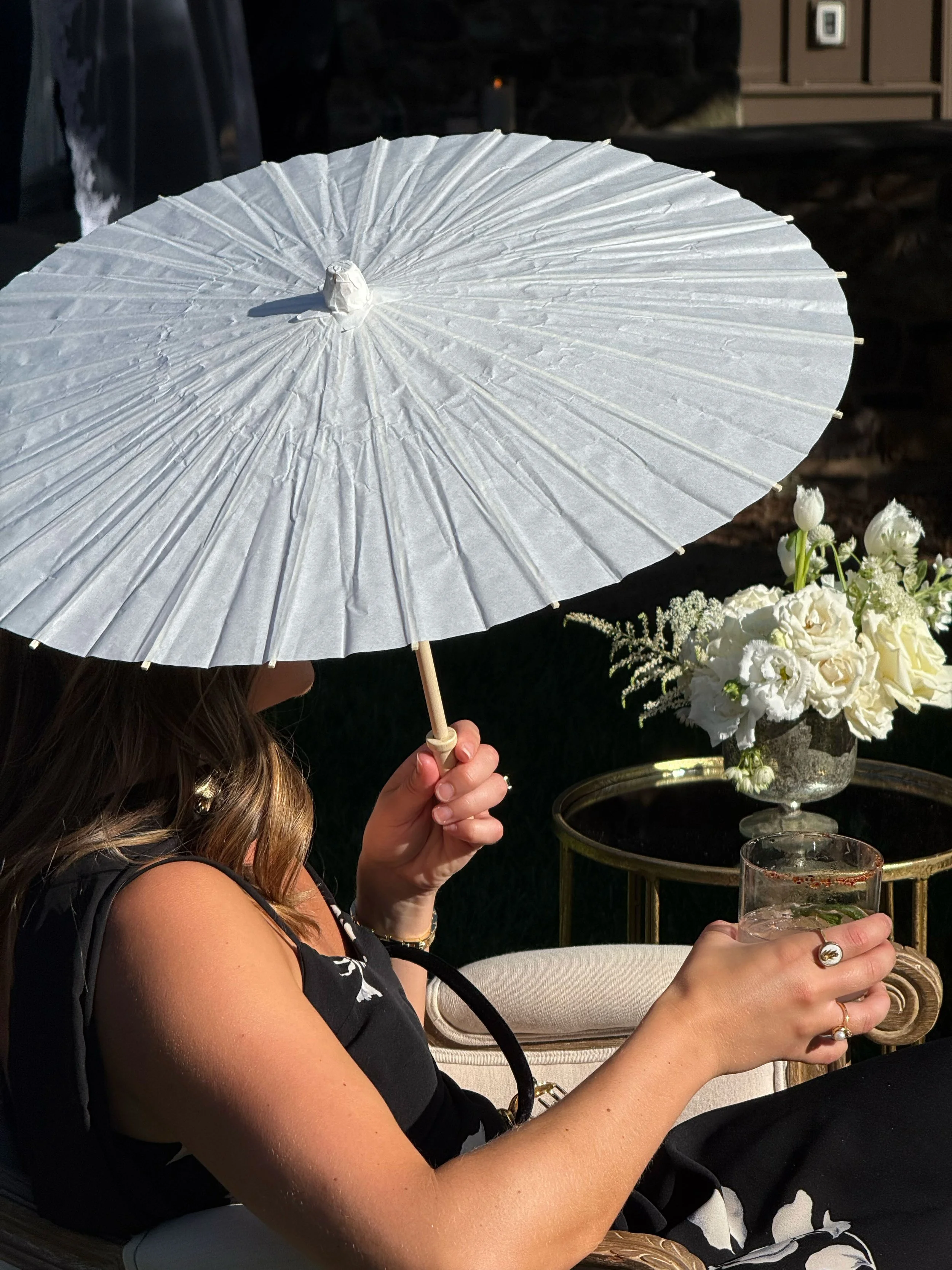 A woman seated outdoors holding a glass of drink, her face hidden behind a large white parasol, with a floral arrangement on a side table in the background.