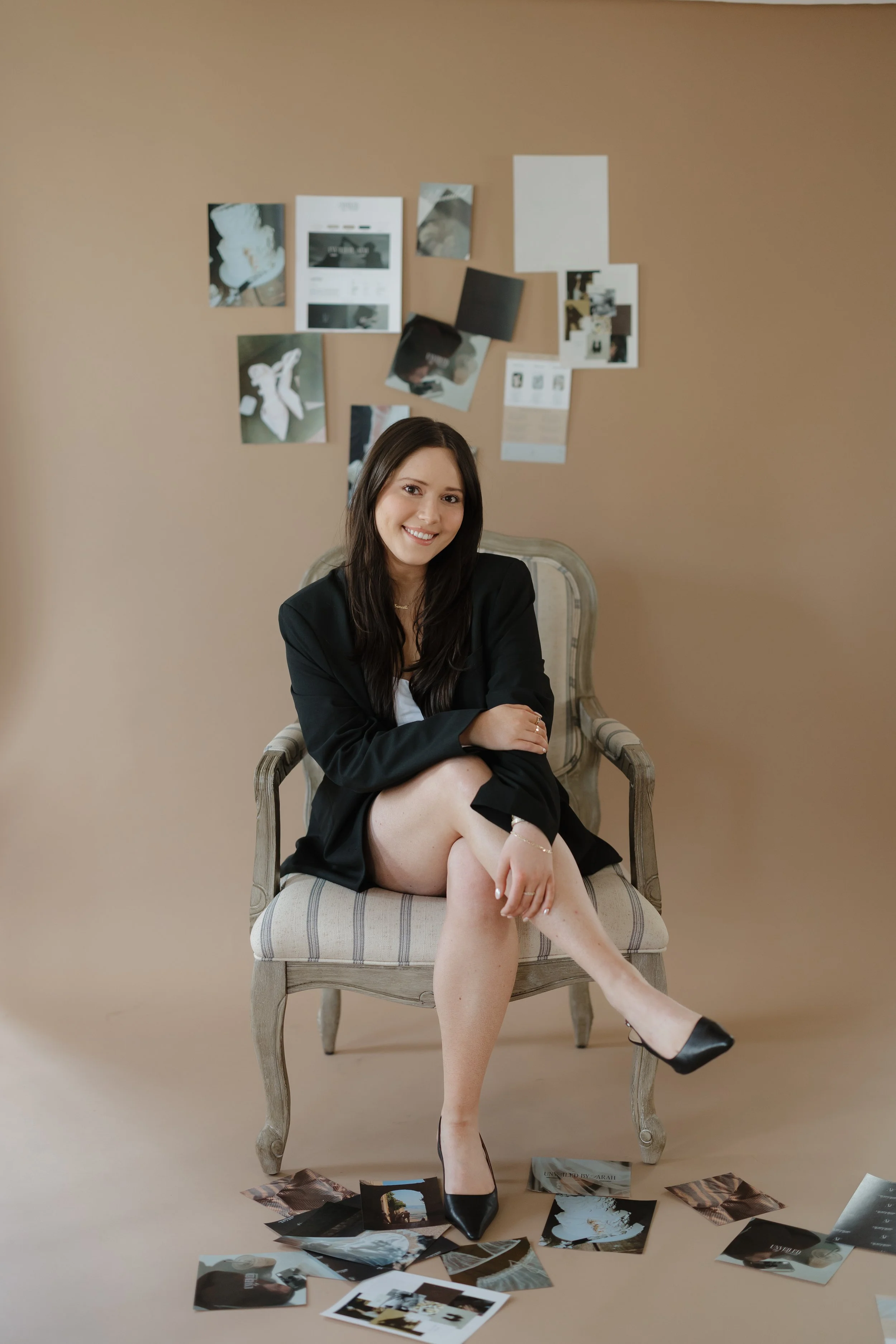 A woman in a black blazer and heels sitting on a vintage armchair with her legs crossed, smiling at the camera, with photographs scattered on the floor and a collage of pictures and notes on the beige wall behind her.