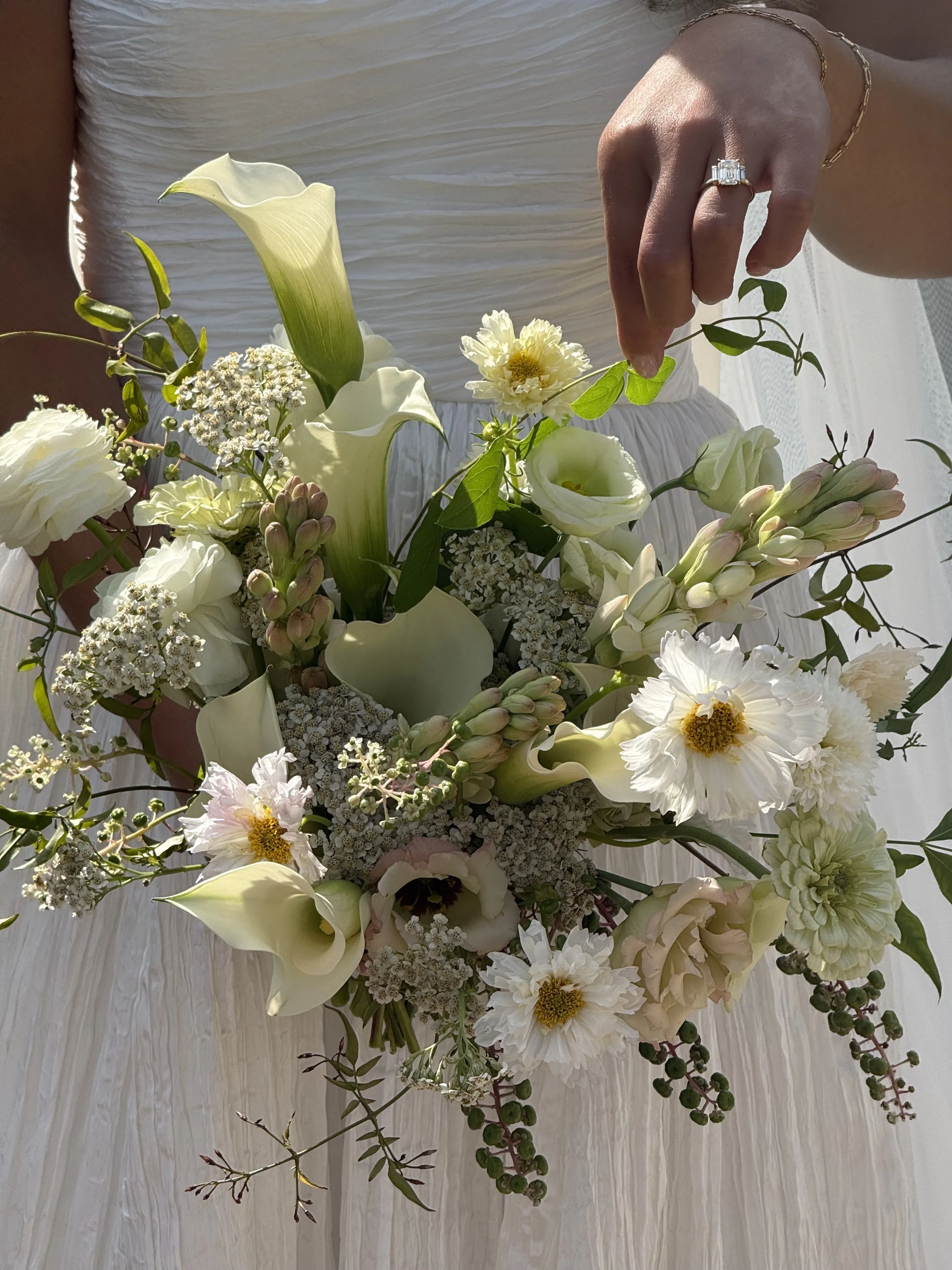 Close-up of a bride holding a bouquet of white and cream flowers, including calla lilies, roses, and daisies, with a wedding dress in the background.