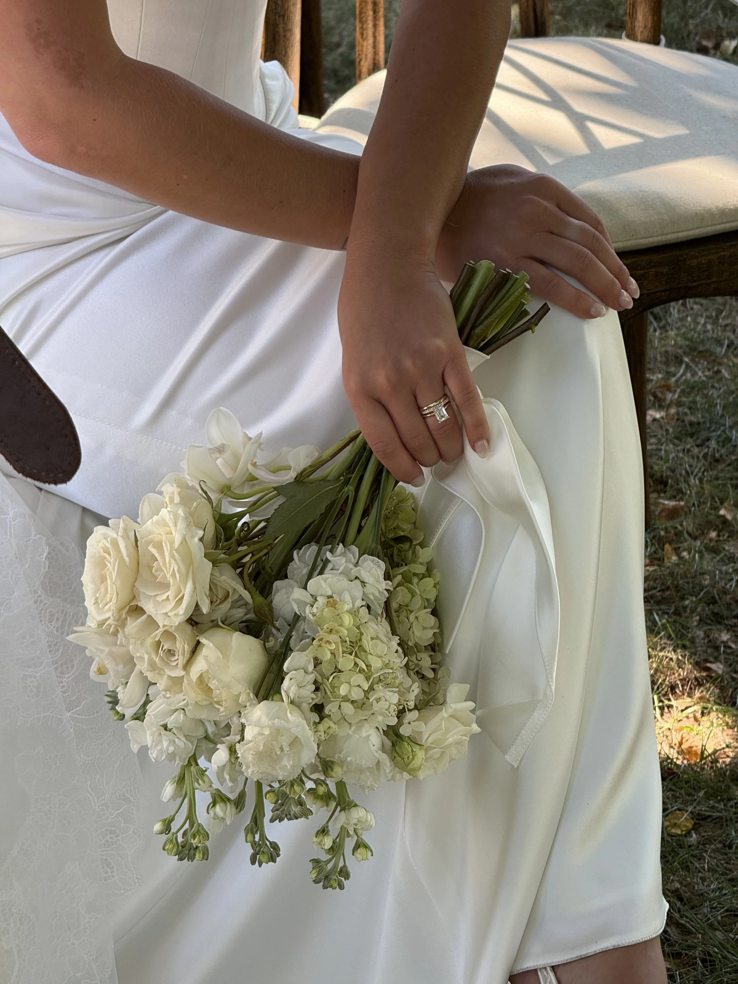 A bride in a white wedding dress is holding a bouquet of white flowers, including roses and hydrangeas, while sitting outdoors.