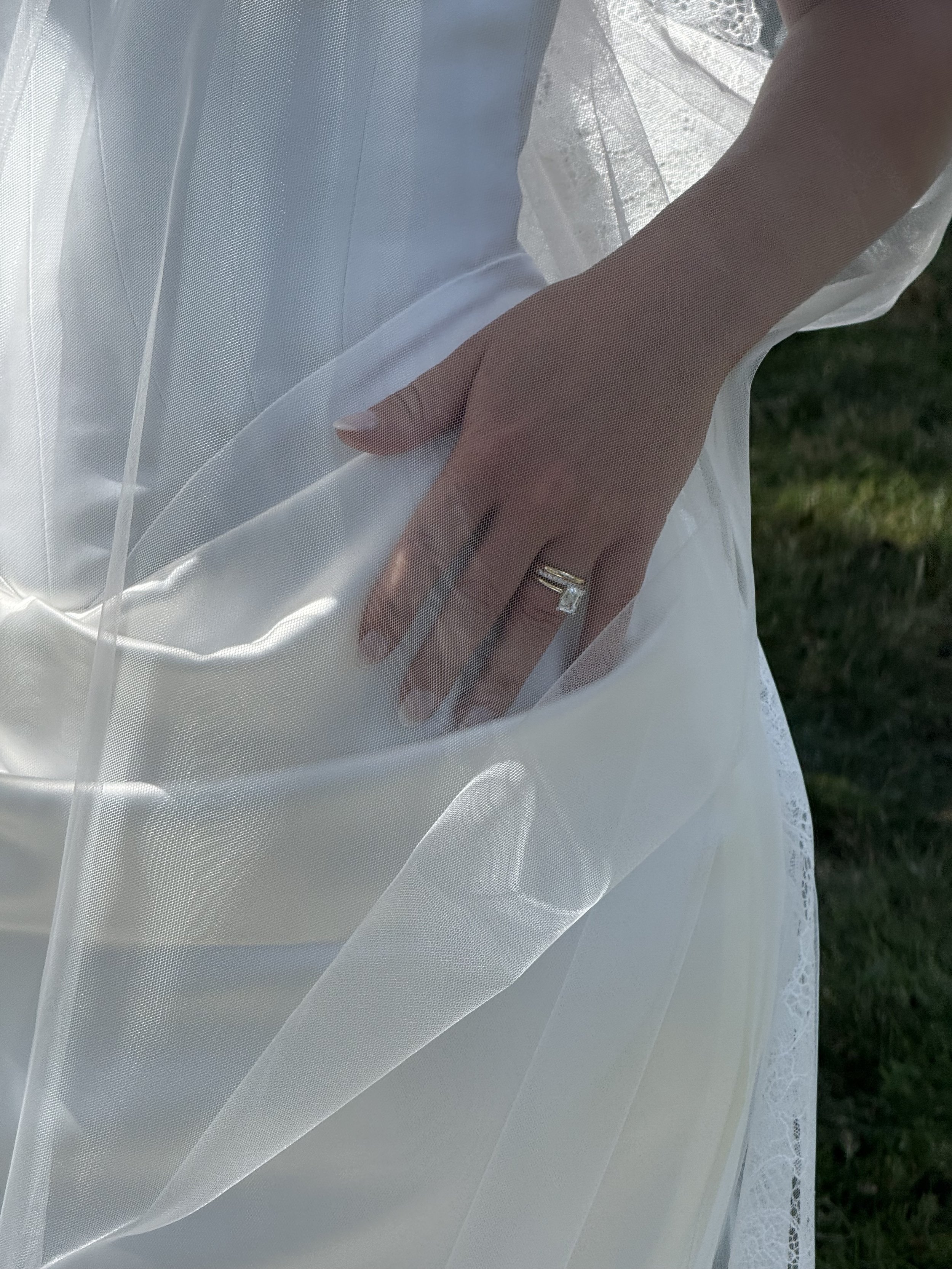 Close-up of a person's hand with a wedding ring on the ring finger, gently pressing on a sheer white veil. The veil is part of a wedding dress, with lace details visible at the edge. The background shows a grassy outdoor setting.