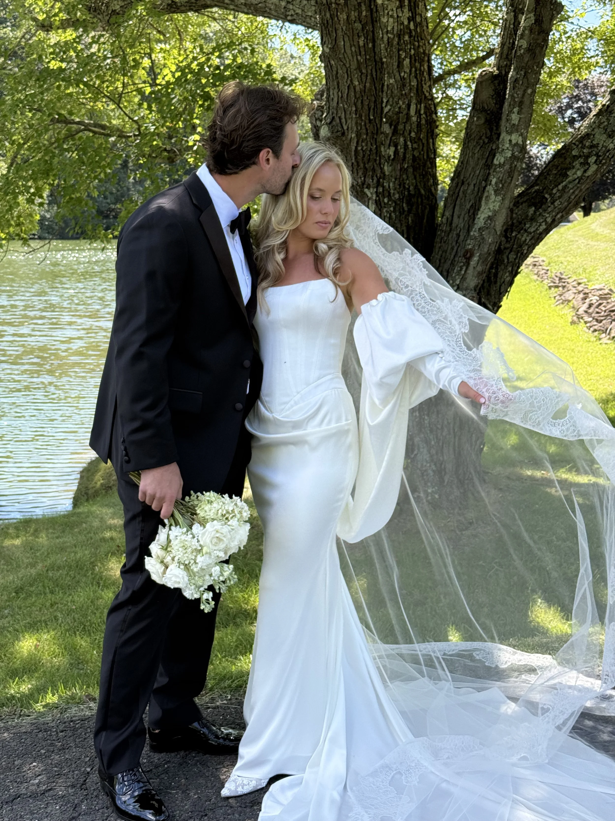 A bride in a white wedding dress and veil stands outdoors near a large tree, holding her veil with one hand, while a groom in a black tuxedo and bowtie kisses her forehead. The groom holds a bouquet of white flowers. There is a body of water and greenery in the background.