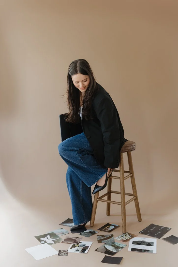 A young woman with long dark hair, wearing a black jacket and blue jeans, sits on a wooden stool holding her black high heel shoe near her foot. Several photos and pieces of paper are scattered on the floor around her against a beige background.