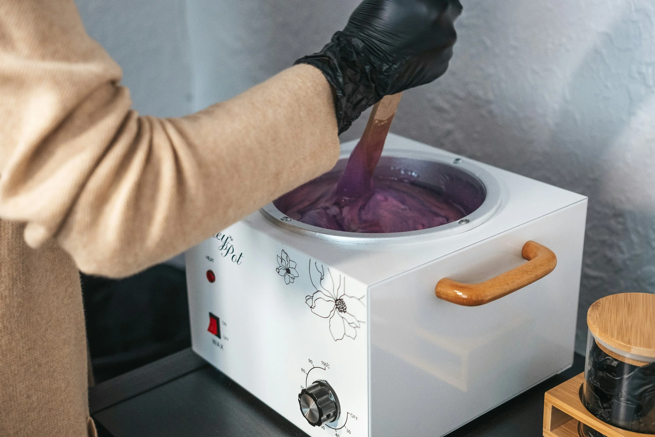 Person stirring a purple substance in a small hot plate with floral design, wearing a beige sweater and black gloves.