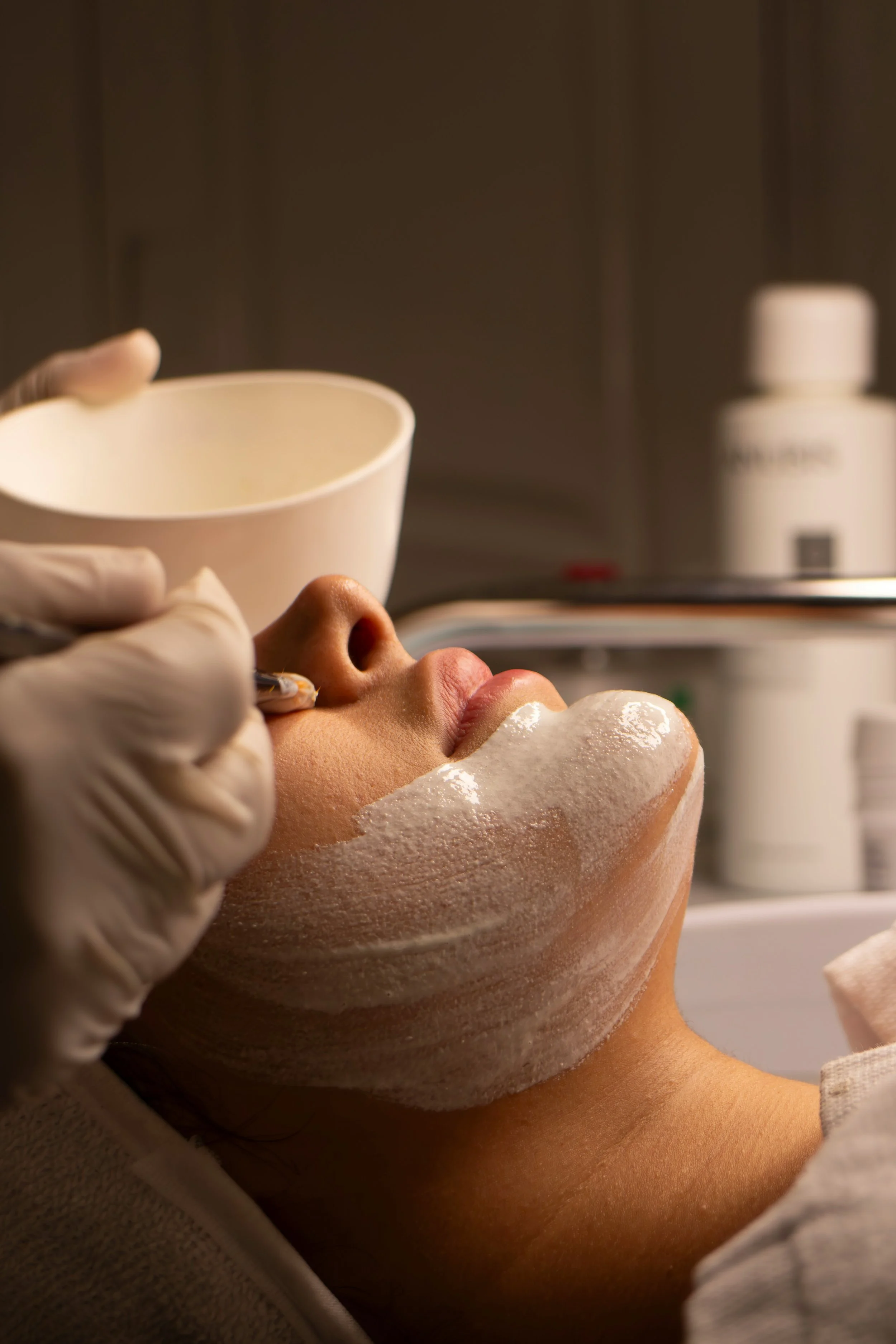 A woman is receiving a facial treatment, lying down with a foam mask on her face. A skincare professional applies a substance with a tool, with skincare products visible in the background.