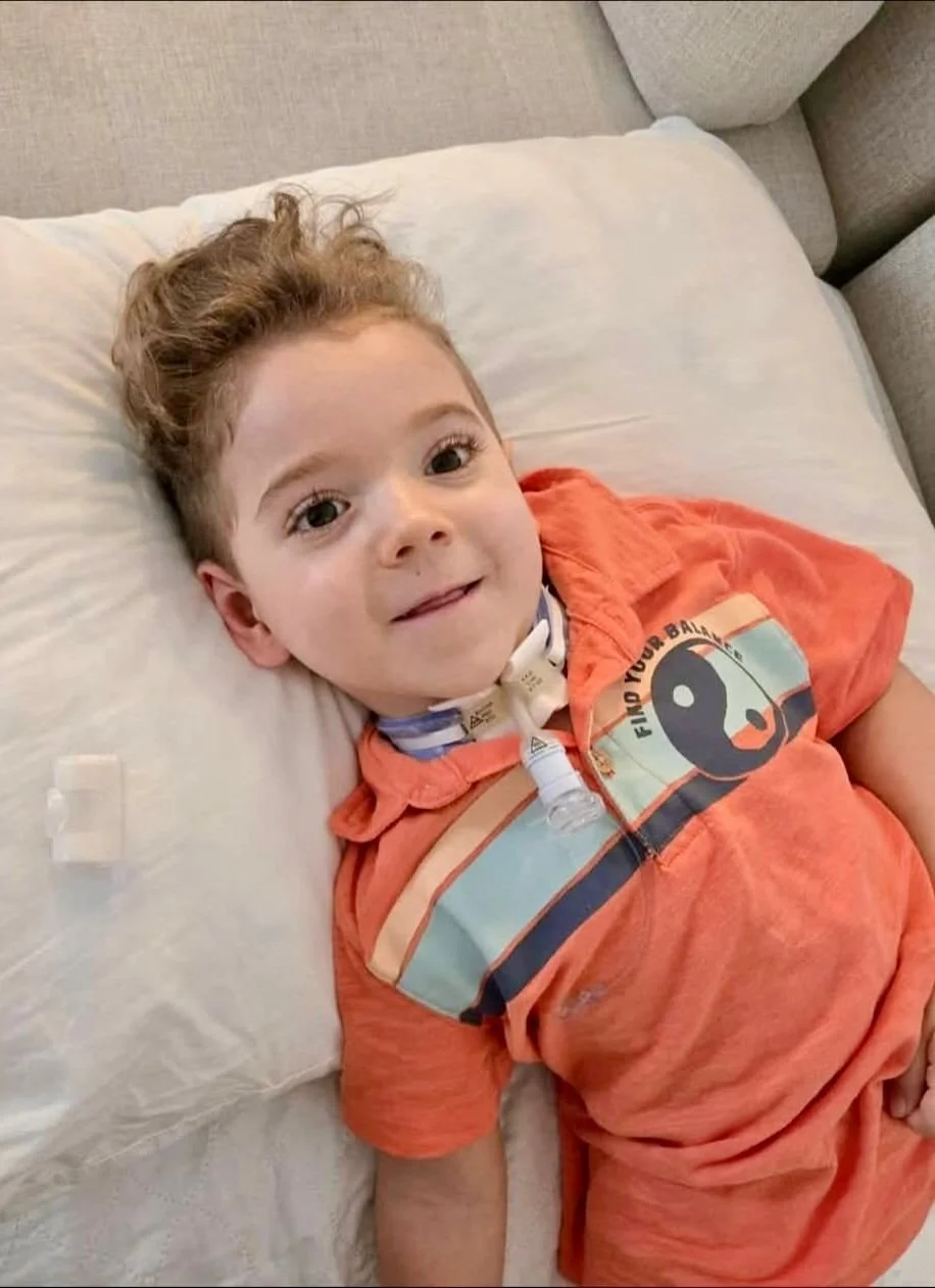 Jeune garçon allongé sur un lit avec un sourire, portant un t-shirt orange avec un dessin de flamant rose et du texte 'Find Your Balance.' Il a un collier et un tube respiratoire autour du cou.