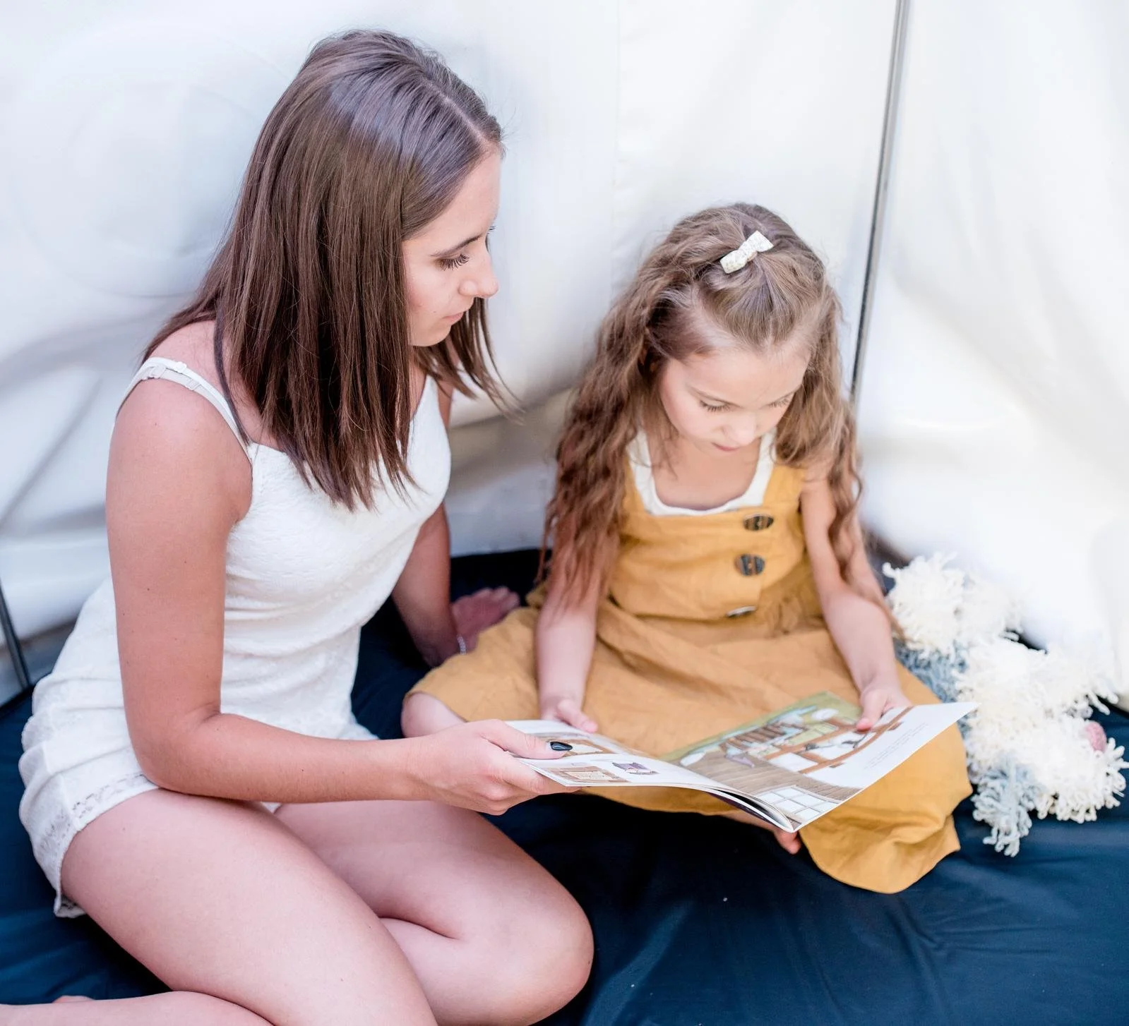 Une femme et une jeune fille regardent un livre ensemble dans un espace blanc et entouré de tissus blancs.