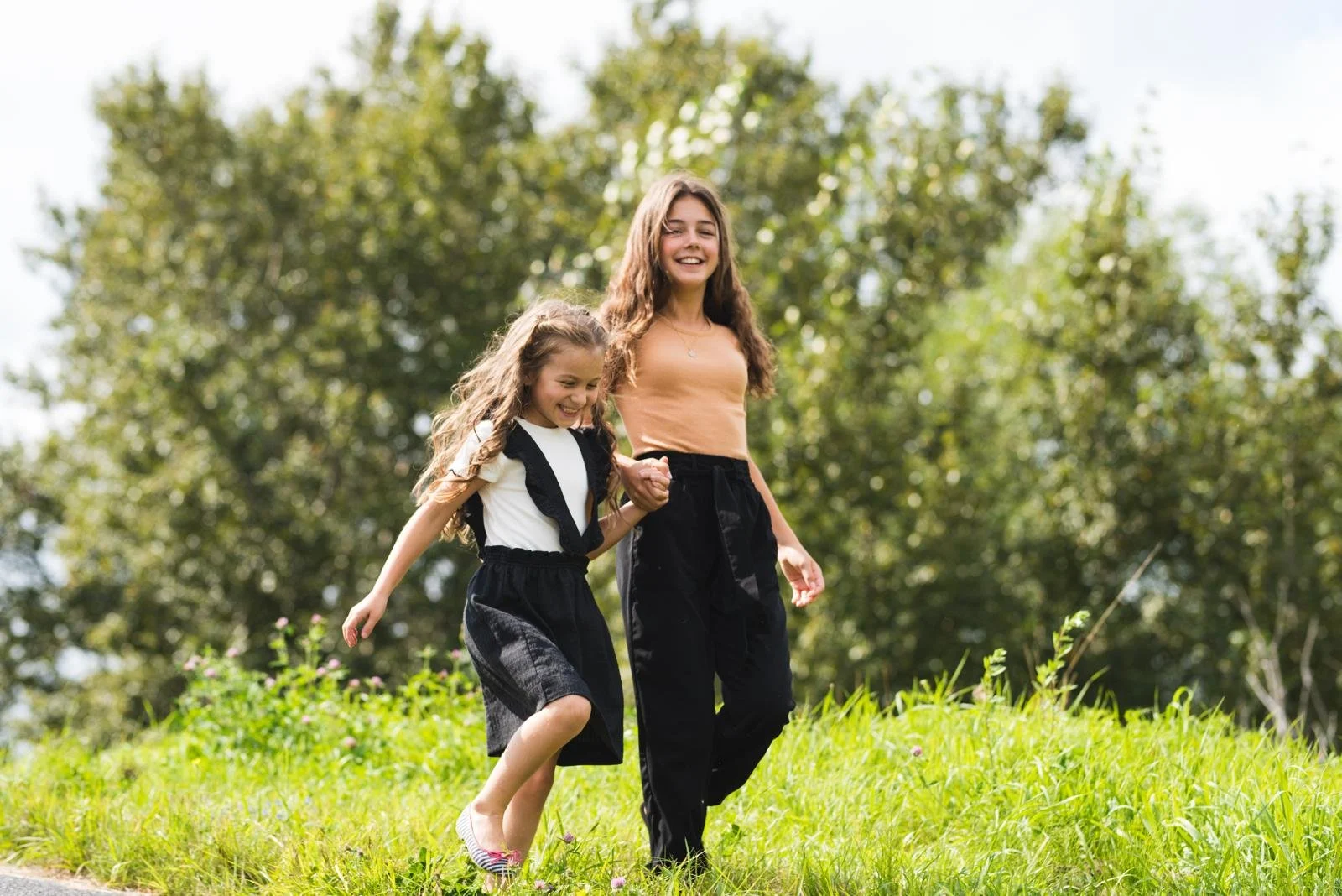 Une jeune femme et une fille marchent main dans la main dans un champ vert avec des arbres en arrière-plan, souriantes et heureuses.