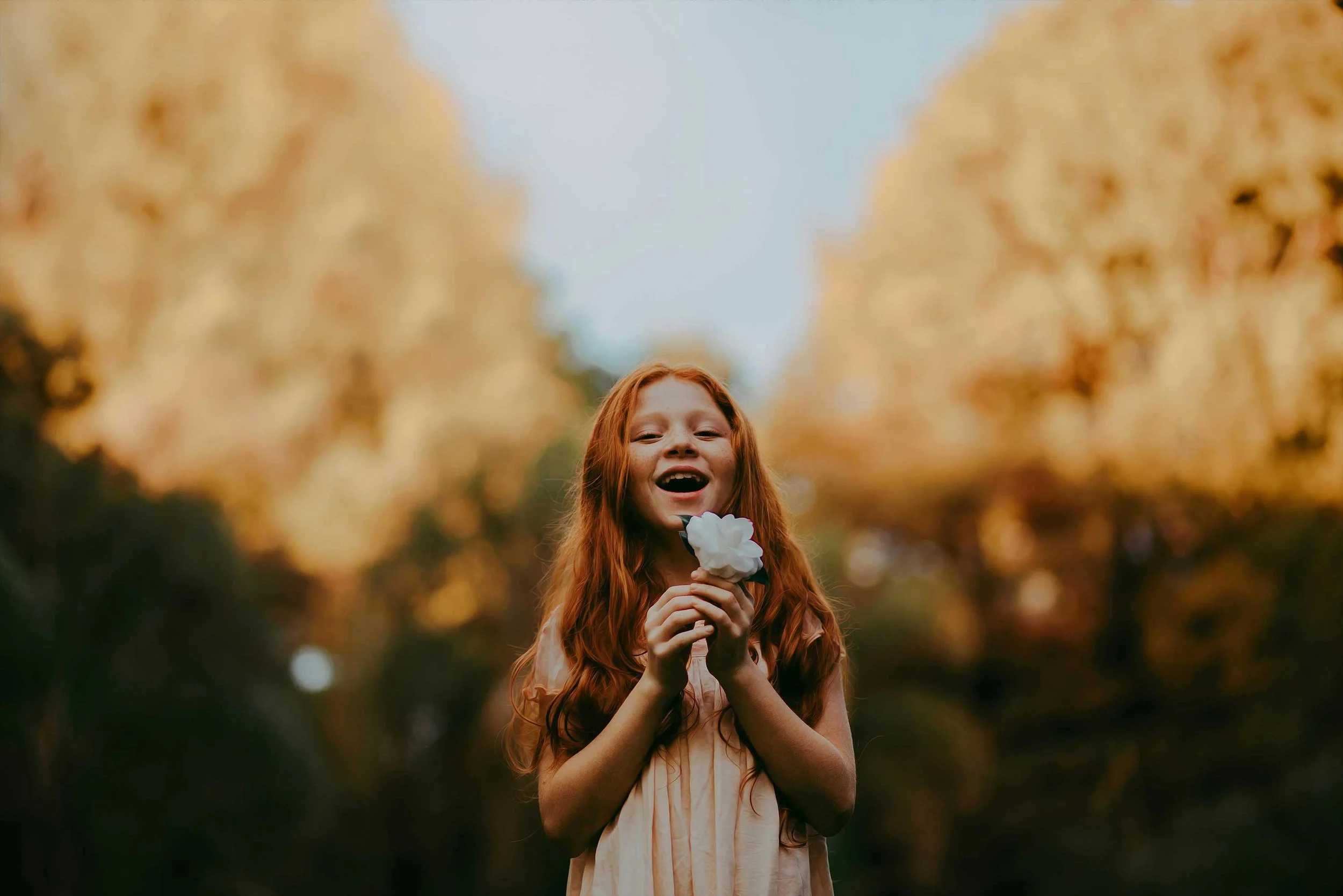 Jeune fille aux cheveux roux, souriante, tenant une fleur blanche devant un fond flou d'arbres en automne.