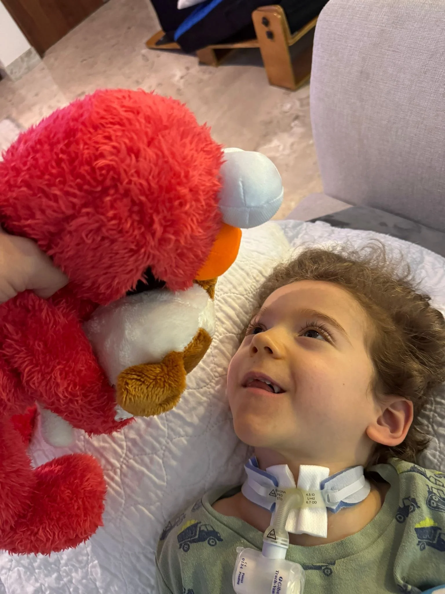 Jeune garçon allongé dans un lit d'hôpital, souriant tout en regardant un animal en peluche rouge, blanc et orange qu'il tient.