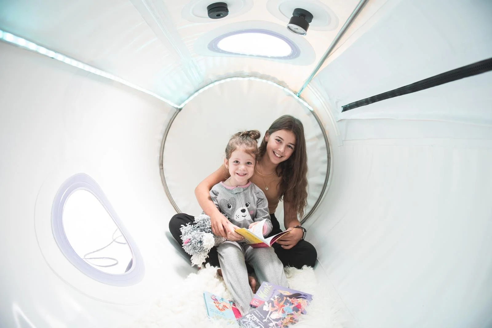 Une jeune femme et une petite fille souriantes assises dans un tunnel de jeu blanc, avec des magazines posés à côté d'elles, partageant un moment de lecture ensemble.