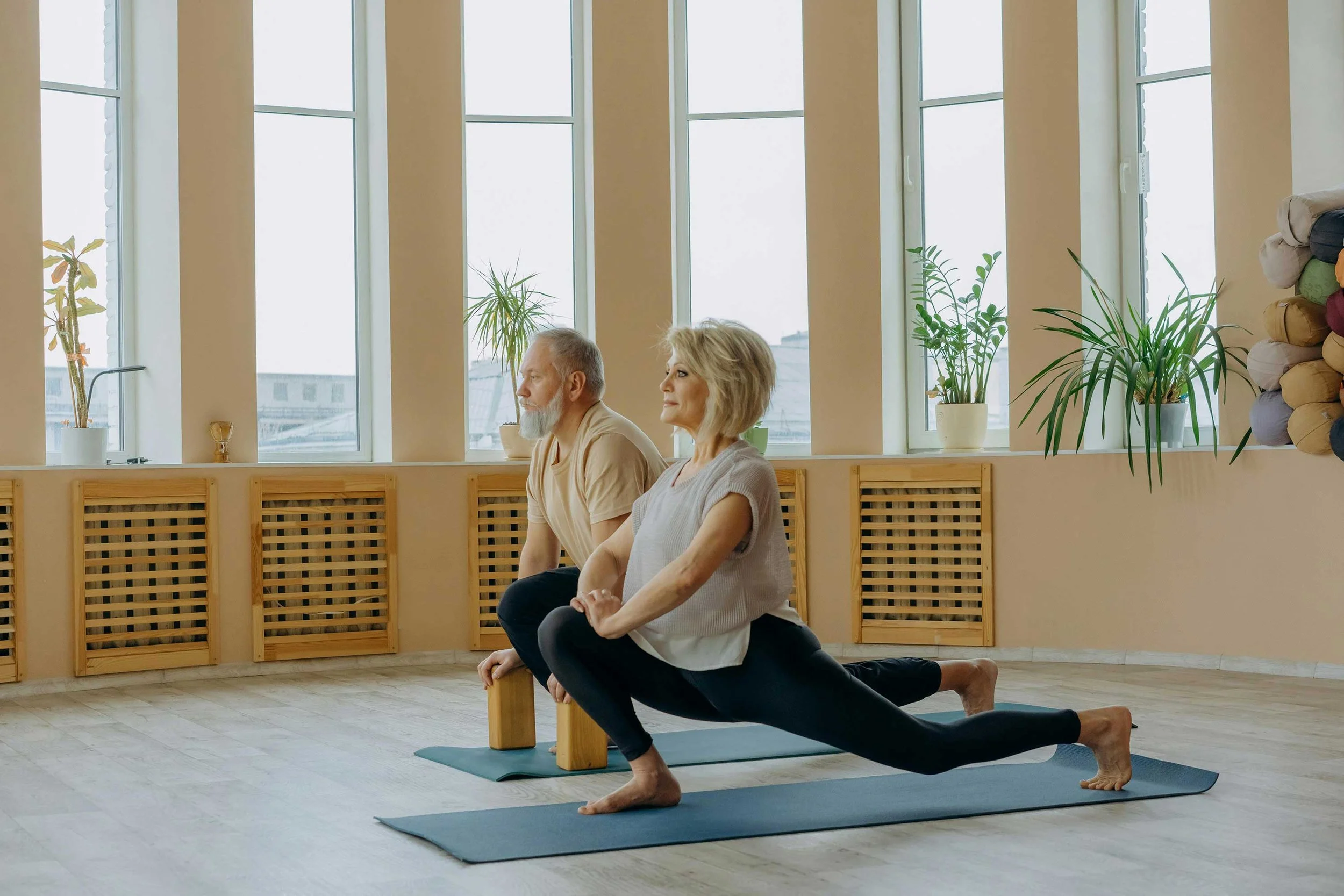 Deux personnes âgées pratiquant le yoga dans une salle lumineuse avec grandes fenêtres et plantes vertes.