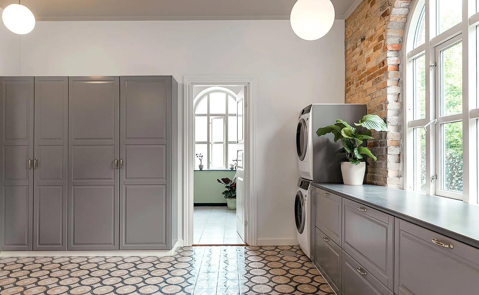 Laundry room with gray cabinets, a washing machine and dryer, a large window, potted plants, and exposed brick wall.