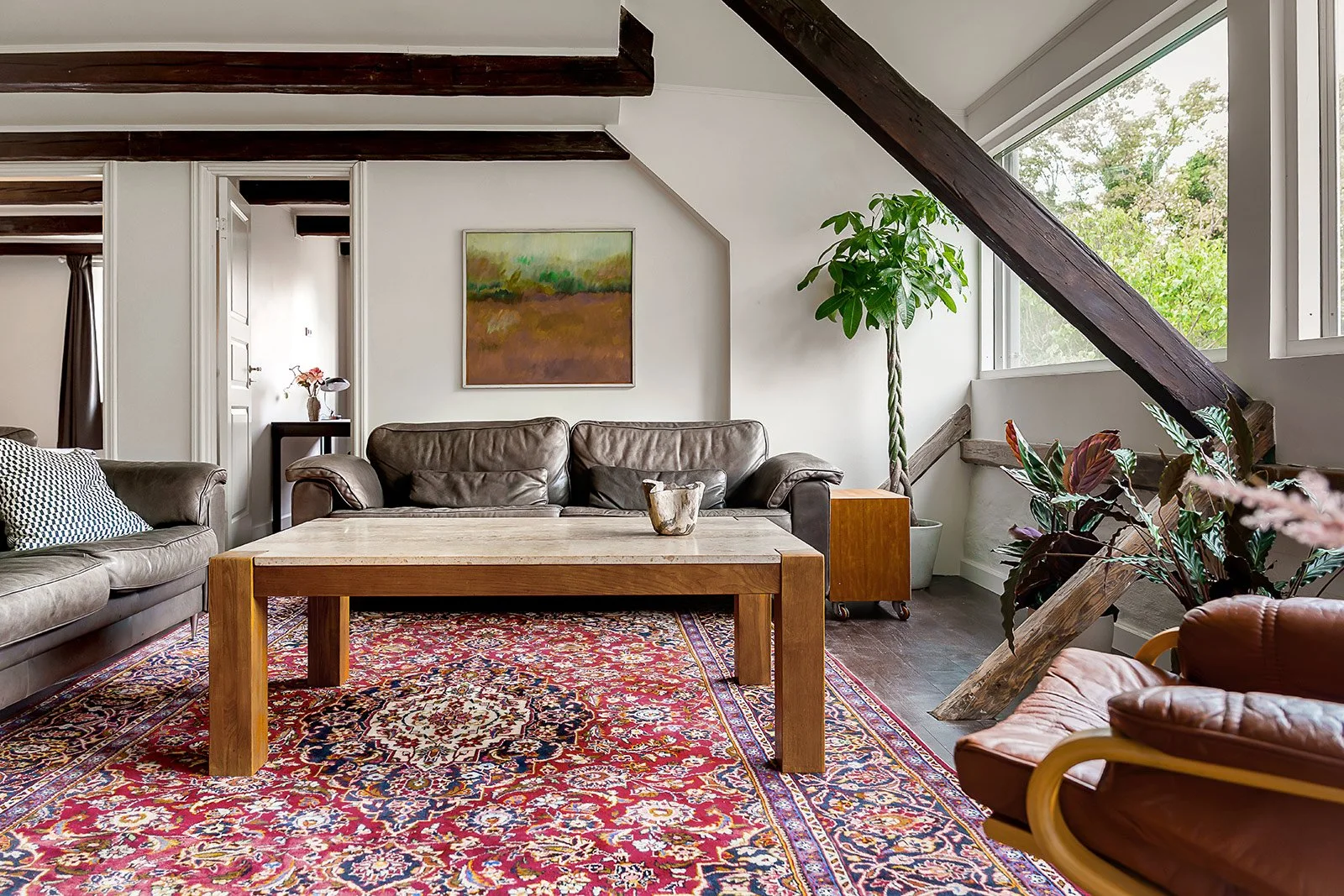 Living room with brown leather sofas, a wooden coffee table, a patterned area rug, a potted plant, and artwork on the wall, with large windows showing greenery outside.