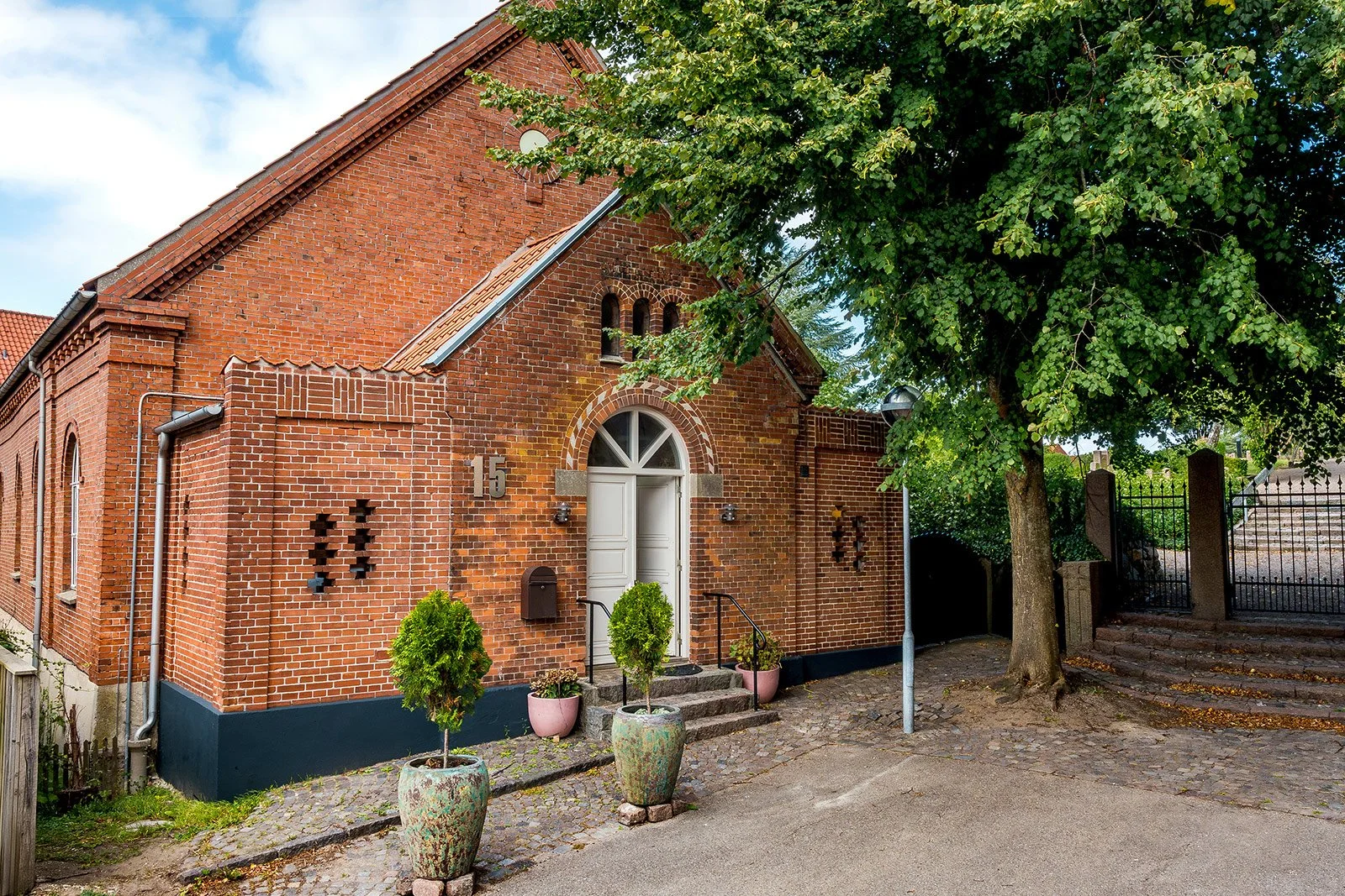 A red brick church with a white door, potted plants on steps, a large tree with green leaves, and a gated pathway on the right.